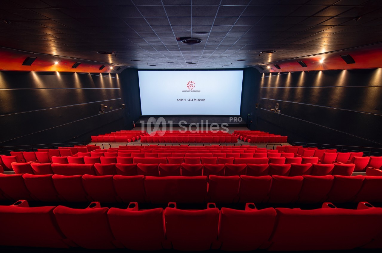 Salle de cinéma vide avec des rangées de sièges rouges, un grand écran blanc et un éclairage tamisé.