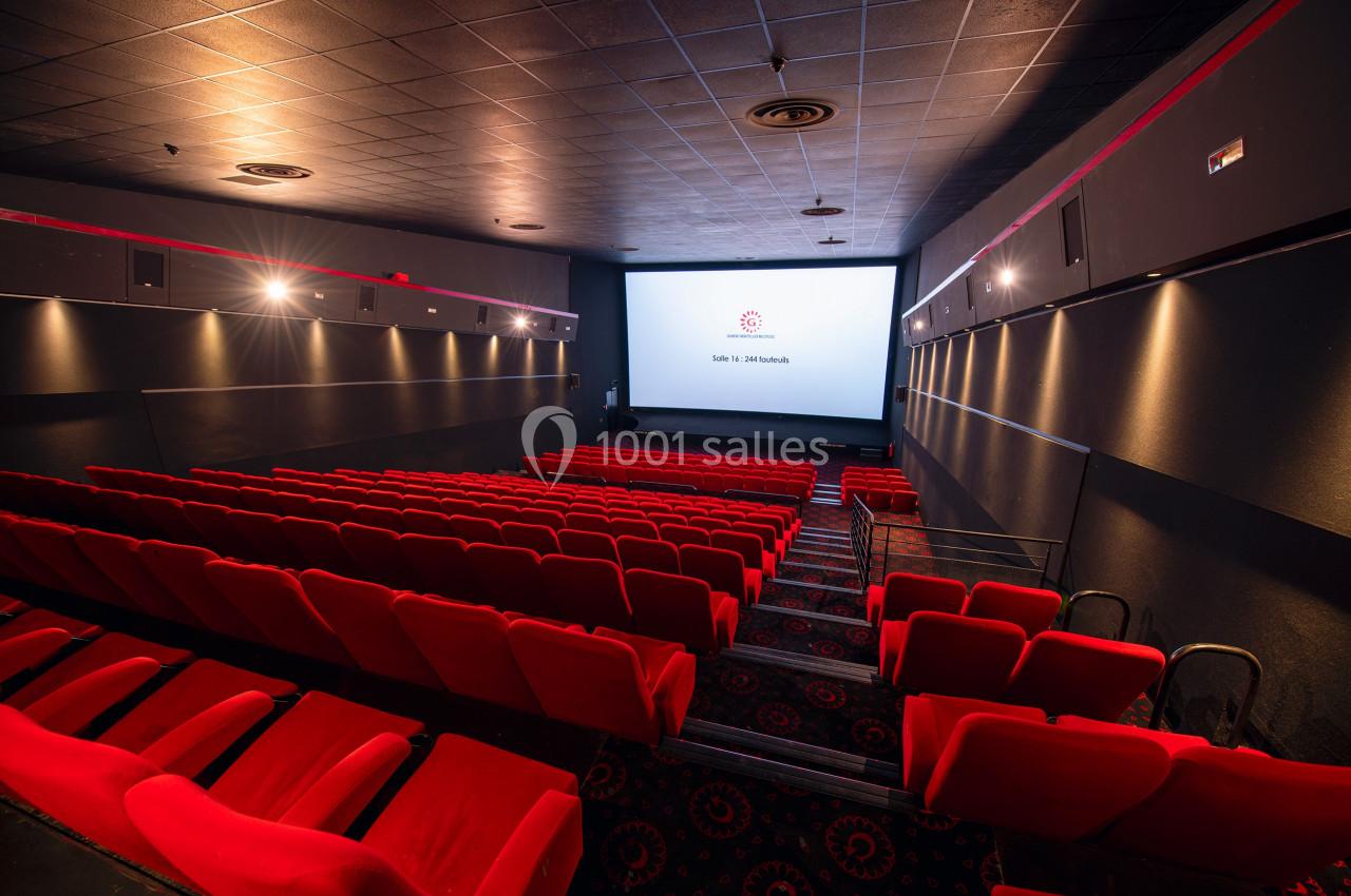Salle de cinéma vide avec des rangées de sièges rouges et un écran blanc allumé au fond.
