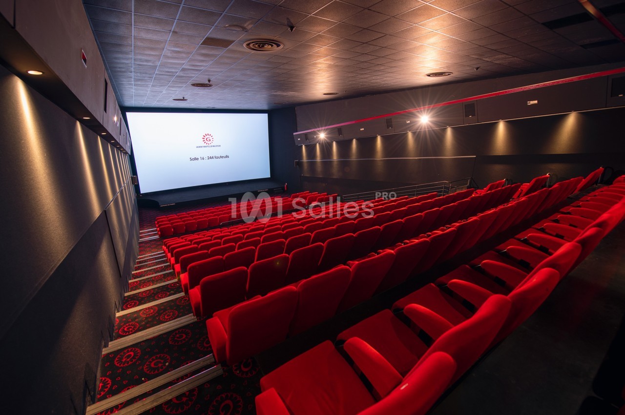 Salle de cinéma vide avec rangées de sièges rouges et écran blanc allumé dans une ambiance tamisée.