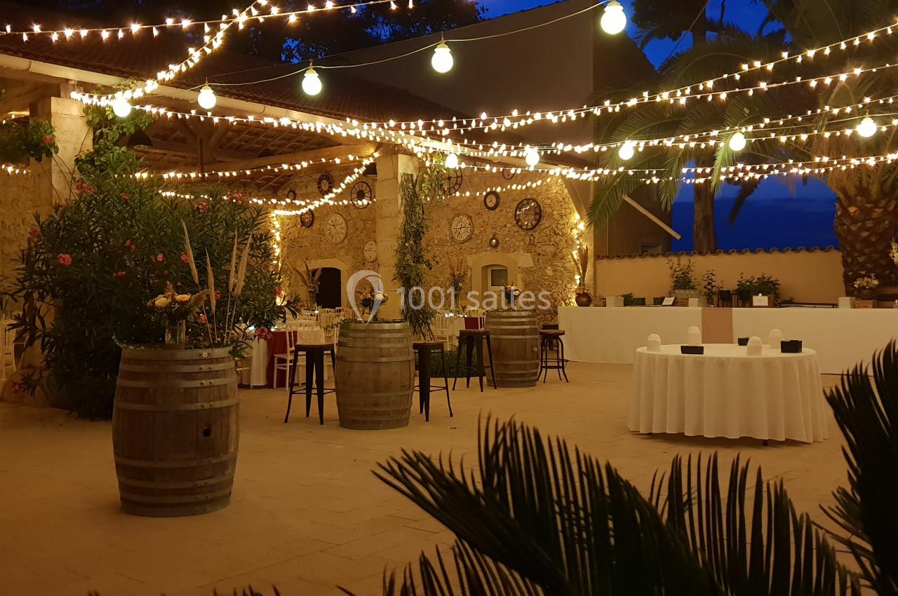 Cour extérieure décorée de guirlandes lumineuses, tables dressées et tonneaux, ambiance festive en soirée.