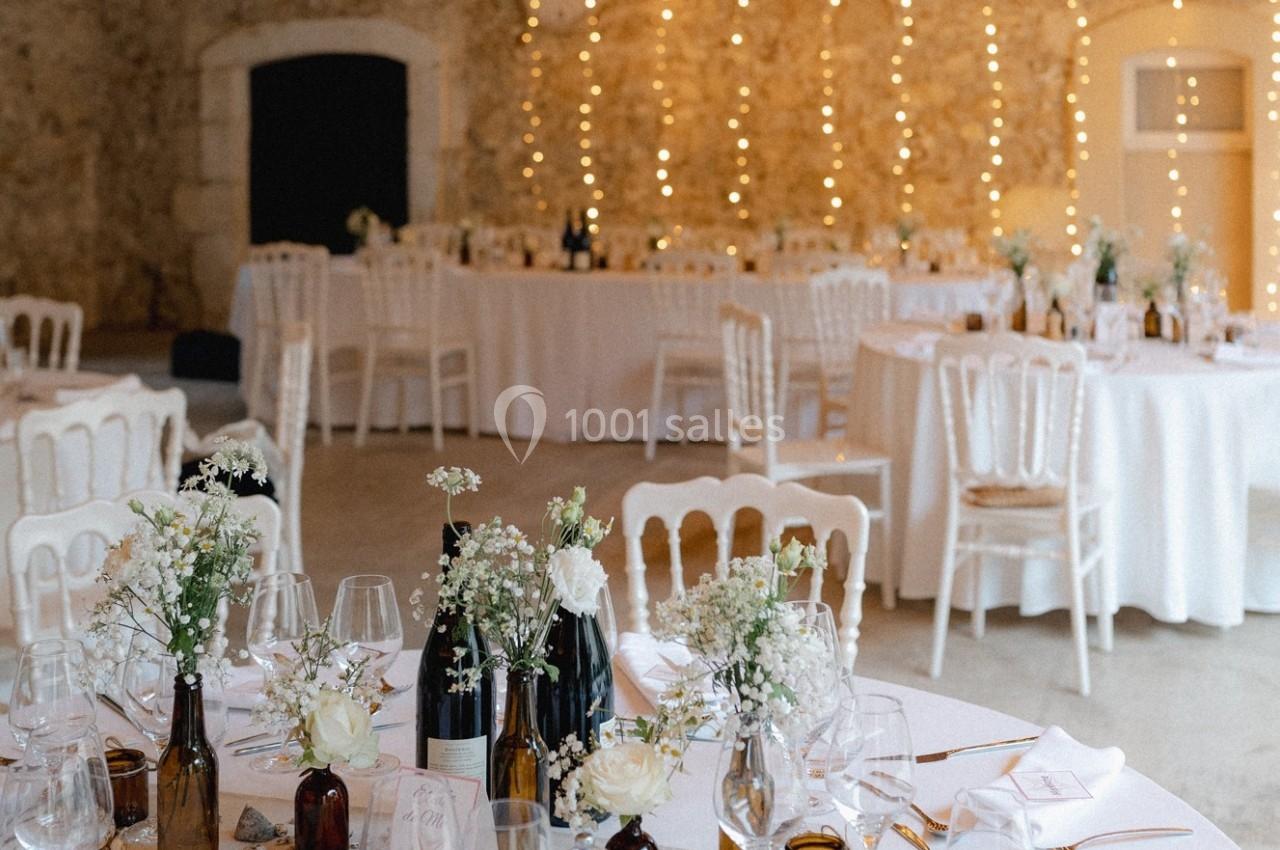 Salle de réception décorée avec des tables rondes, nappes blanches, chaises assorties et guirlandes lumineuses en arrière…