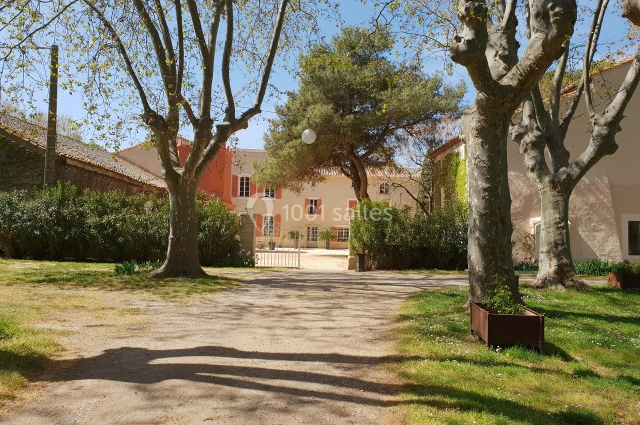 Allée bordée d'arbres menant à une maison aux façades claires, entourée de végétation sous un ciel dégagé.