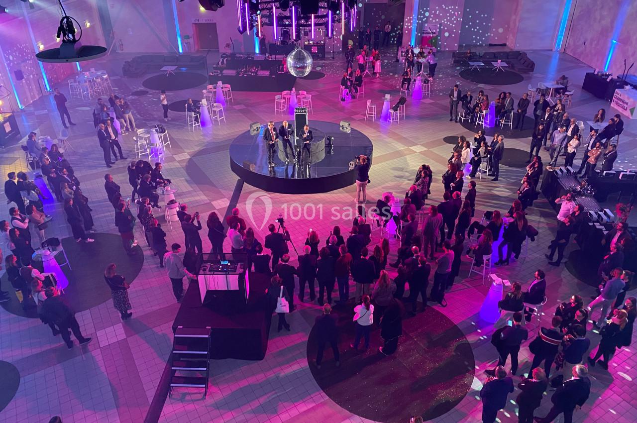 Vue aérienne d'un événement en intérieur avec des invités rassemblés autour d'une scène centrale sous une lumière colorée.