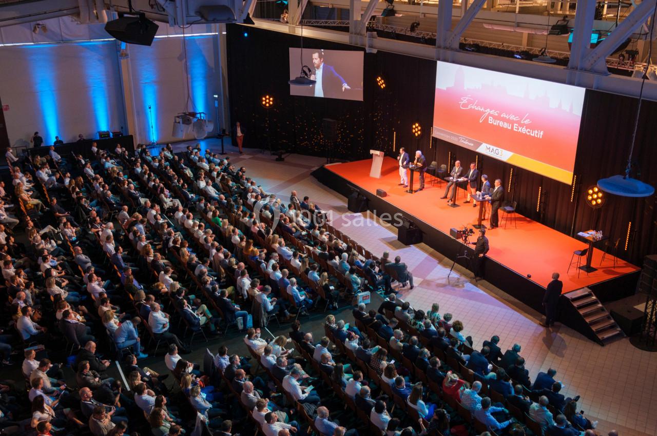 Vue d'une conférence avec un public nombreux, une scène éclairée et des intervenants discutant sur un podium.