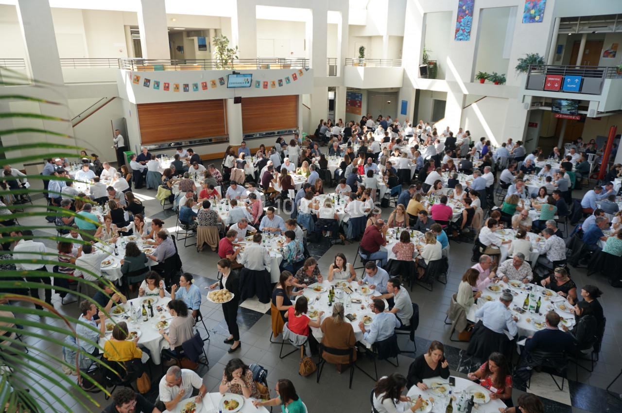 Salle lumineuse remplie de nombreuses tables rondes où des personnes partagent un repas dans une ambiance conviviale.