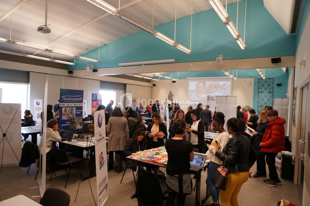 Salle animée avec des stands d'information, des visiteurs échangeant et un écran projetant une présentation.