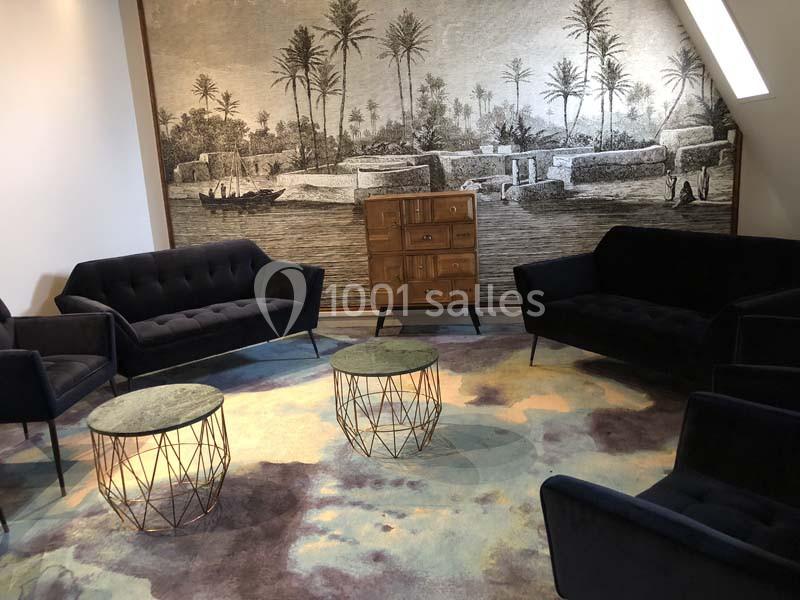 Salon avec canapés noirs, tables basses rondes, tapis coloré et fresque murale représentant un paysage exotique.