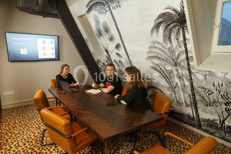 Trois personnes discutent autour d'une table en bois dans une salle de réunion au décor tropical.