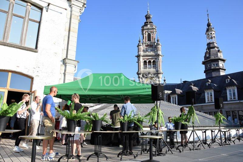 Personnes rassemblées sous une tente verte sur une terrasse, avec des plantes en pot et des clochers en arrière-plan.