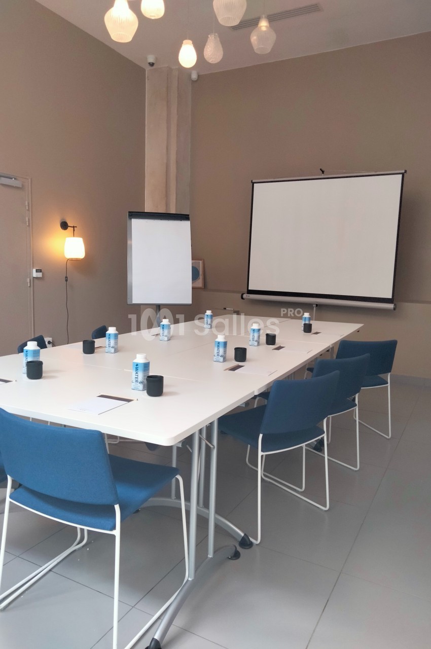 Salle de réunion avec table rectangulaire, chaises bleues, paperboard, vidéoprojecteur et bouteilles d'eau disposées.