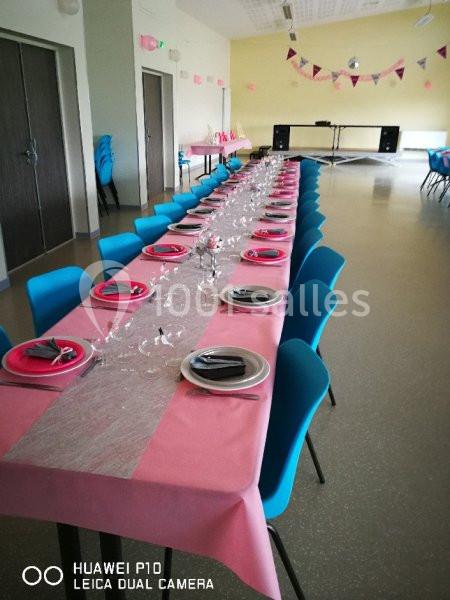 Table dressée avec nappes roses, assiettes et couverts, dans une salle décorée avec guirlandes et chaises bleues.