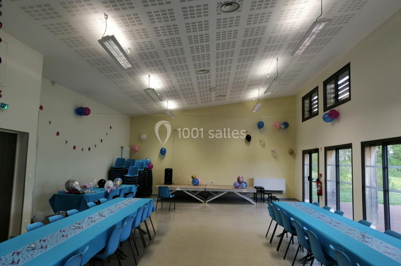 Salle décorée pour un événement avec tables alignées, nappes bleues, ballons et chaises empilées au fond.