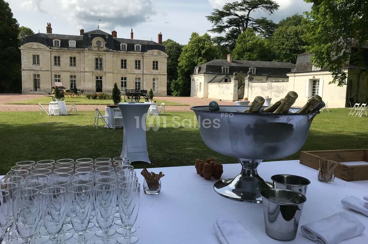 Table dressée avec des flûtes et un seau à champagne devant un château entouré de verdure par temps ensoleillé.