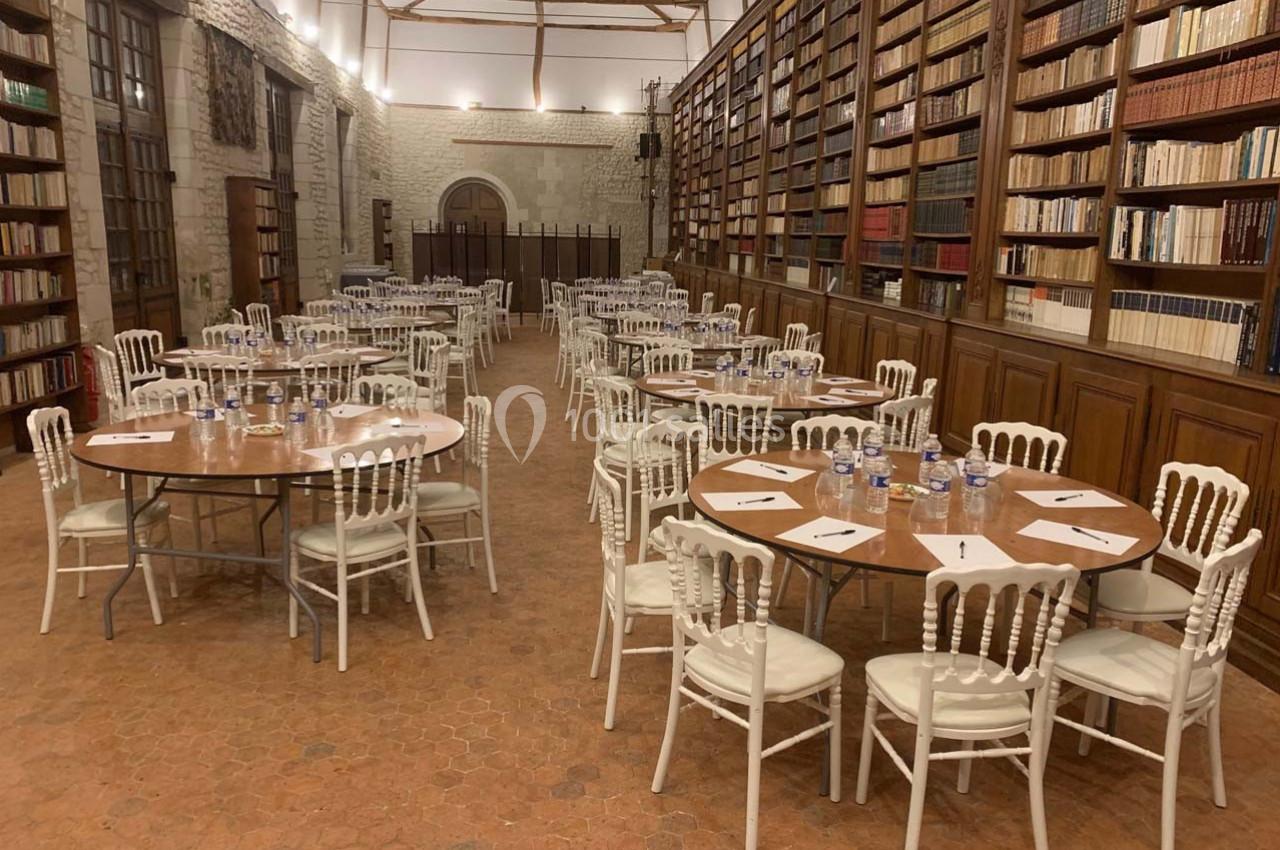 Salle avec des tables rondes dressées, chaises blanches et étagères remplies de livres le long des murs.