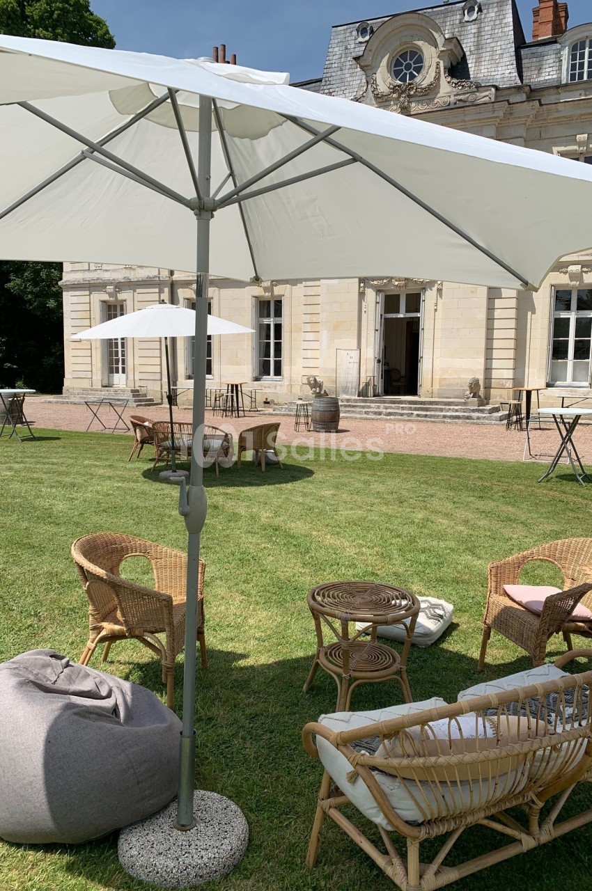 Mobilier de jardin en rotin disposé sous un parasol blanc sur une pelouse devant un bâtiment en pierre.