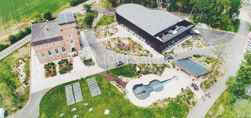 Vue aérienne d'un domaine avec un bâtiment en forme de guitare, un manoir en briques et des jardins aménagés.