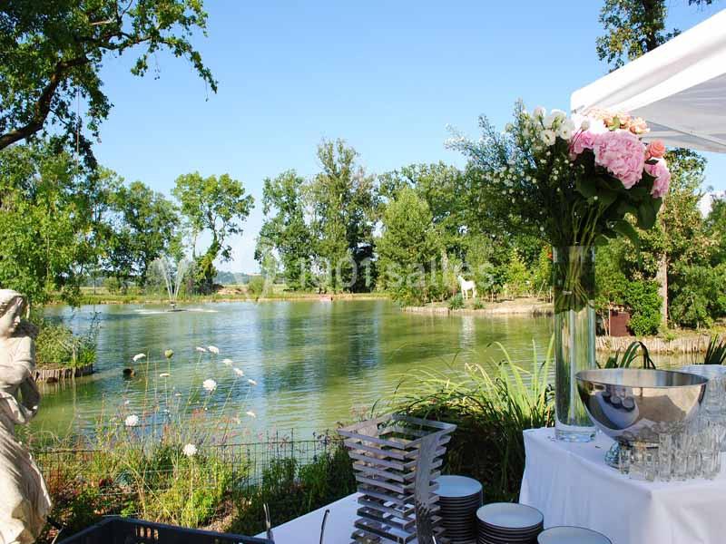 Vue d'un lac entouré de verdure avec une table dressée en premier plan, décorée de fleurs et d'ustensiles.
