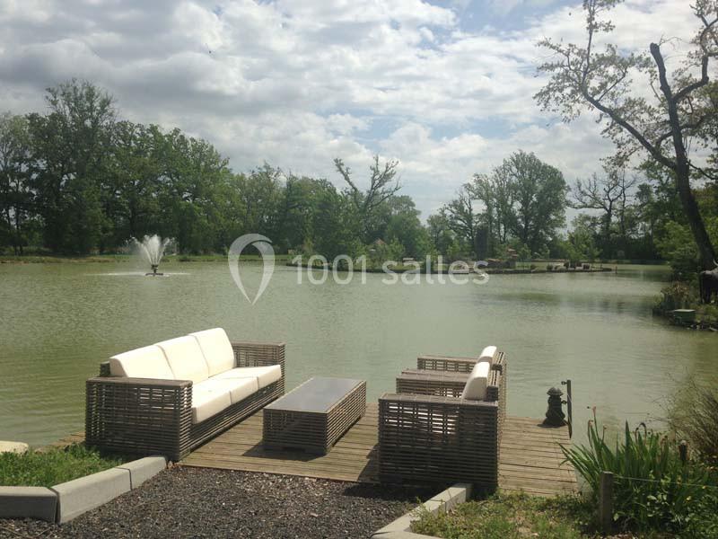 Mobilier d'extérieur en rotin installé sur une terrasse en bois au bord d'un étang avec une fontaine et des arbres en…