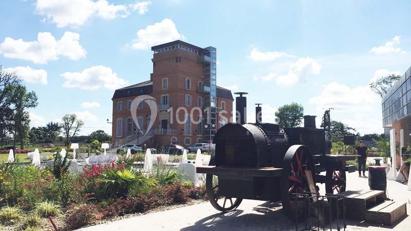 Ancienne locomotive exposée devant un bâtiment en briques entouré de jardins fleuris sous un ciel ensoleillé.