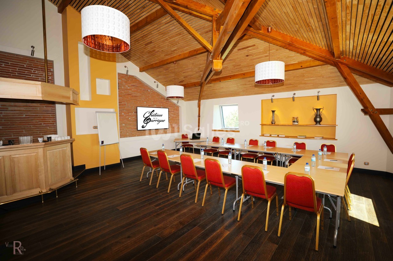 Salle de réunion lumineuse avec poutres apparentes, tables disposées en U, chaises rouges et écran mural.