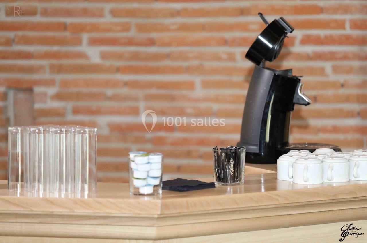 Machine à café noire sur un comptoir en bois avec des tasses blanches, des capsules et des verres transparents alignés.
