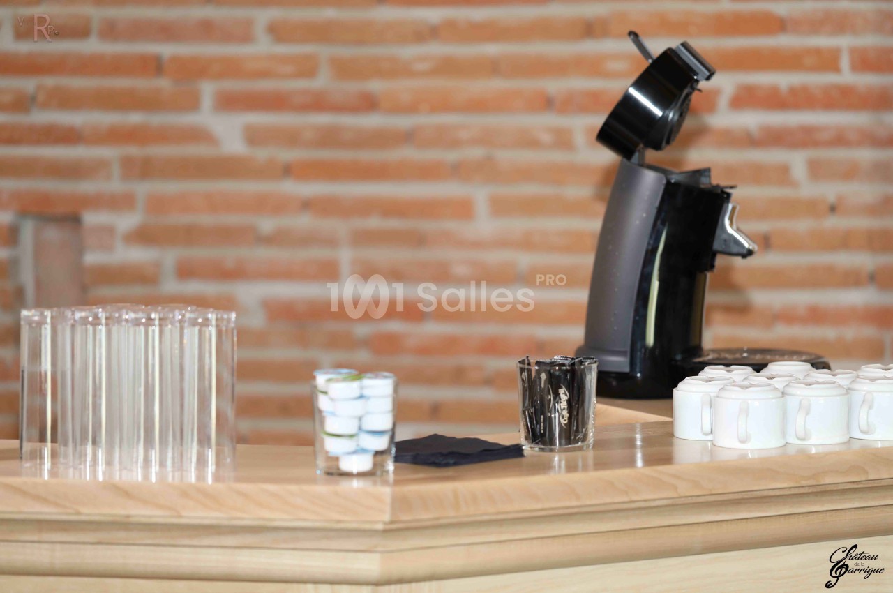 Machine à café noire sur un comptoir en bois avec des tasses blanches, des capsules et des verres transparents alignés.