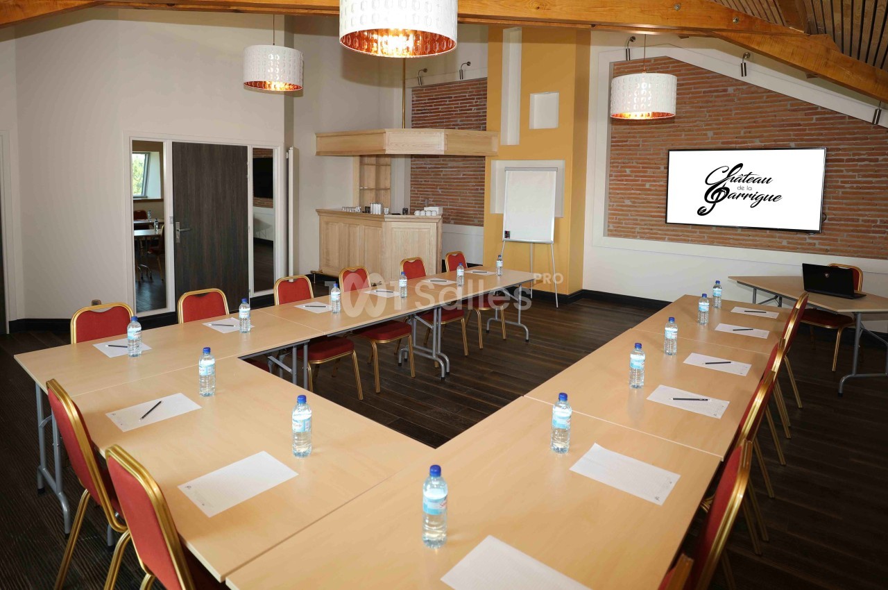 Salle de réunion lumineuse avec tables disposées en U, chaises rouges, bouteilles d'eau et écran de présentation.