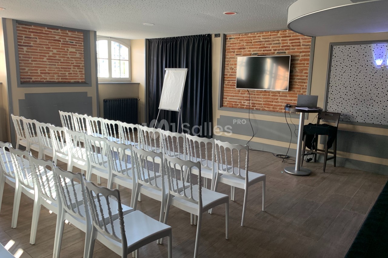 Salle de réunion lumineuse avec chaises blanches alignées, écran, chevalet de présentation et murs en briques apparentes.