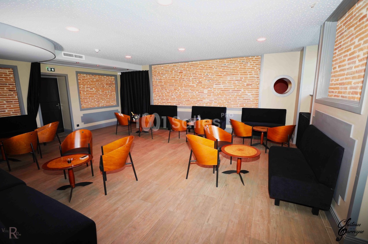 Salle avec murs en briques, mobilier en bois et fauteuils orange, disposés autour de petites tables rondes.