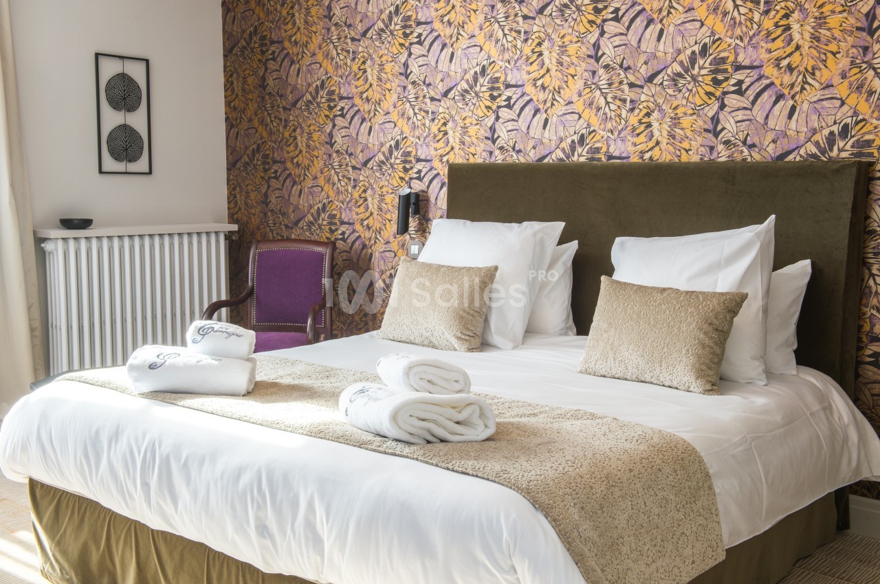 Lit double avec coussins et serviettes pliées, devant un mur à motifs floraux et une chaise violette.