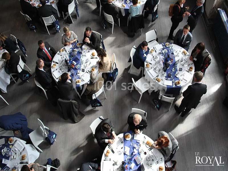 Vue aérienne d'invités assis autour de tables rondes dressées pour un repas dans une salle lumineuse.