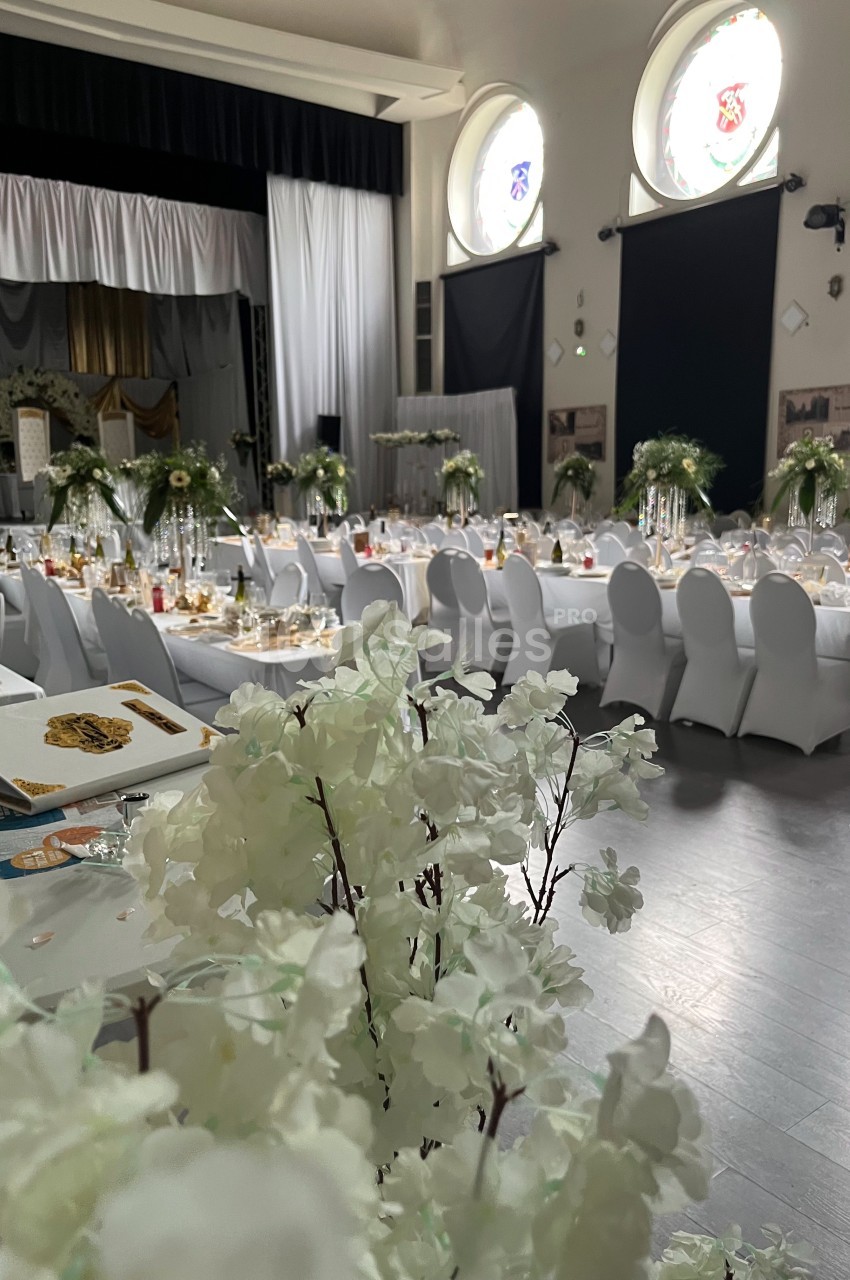 Salle de réception décorée pour un événement, avec tables dressées, chaises blanches et arrangements floraux élégants.