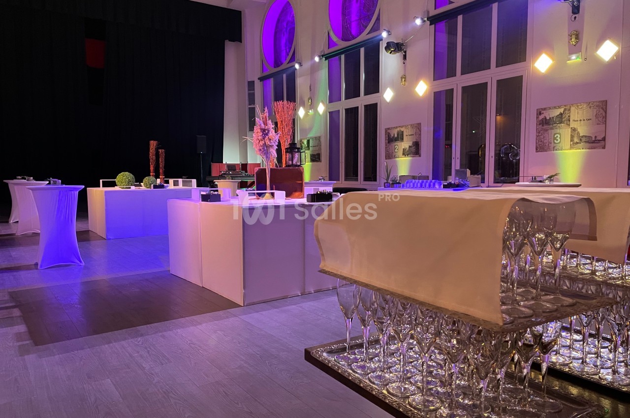 Salle de réception élégamment éclairée avec tables hautes, décorations modernes et verres disposés sur un chariot.