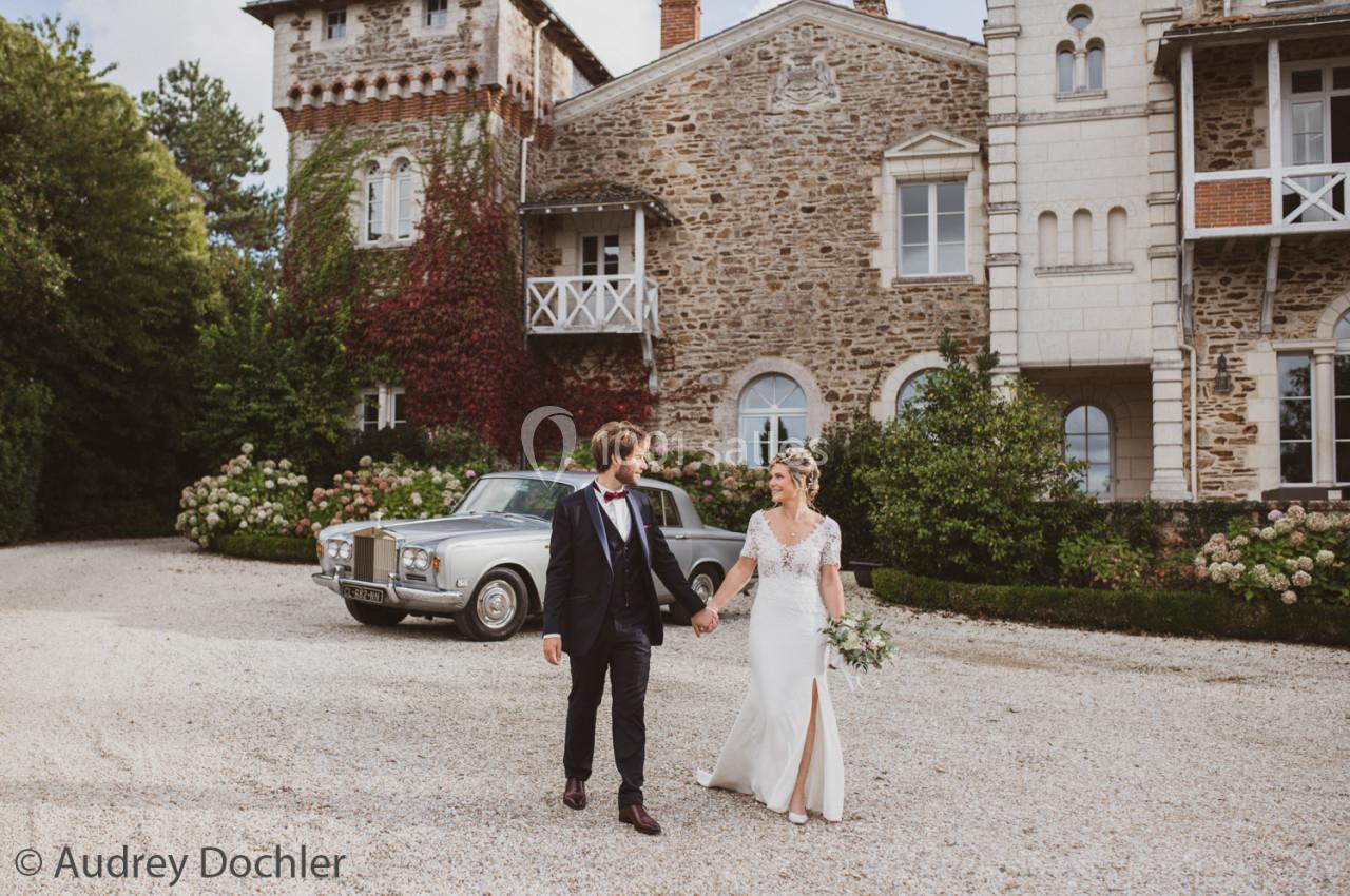 Un couple en tenue de mariage marche main dans la main devant un manoir en pierre et une voiture ancienne.