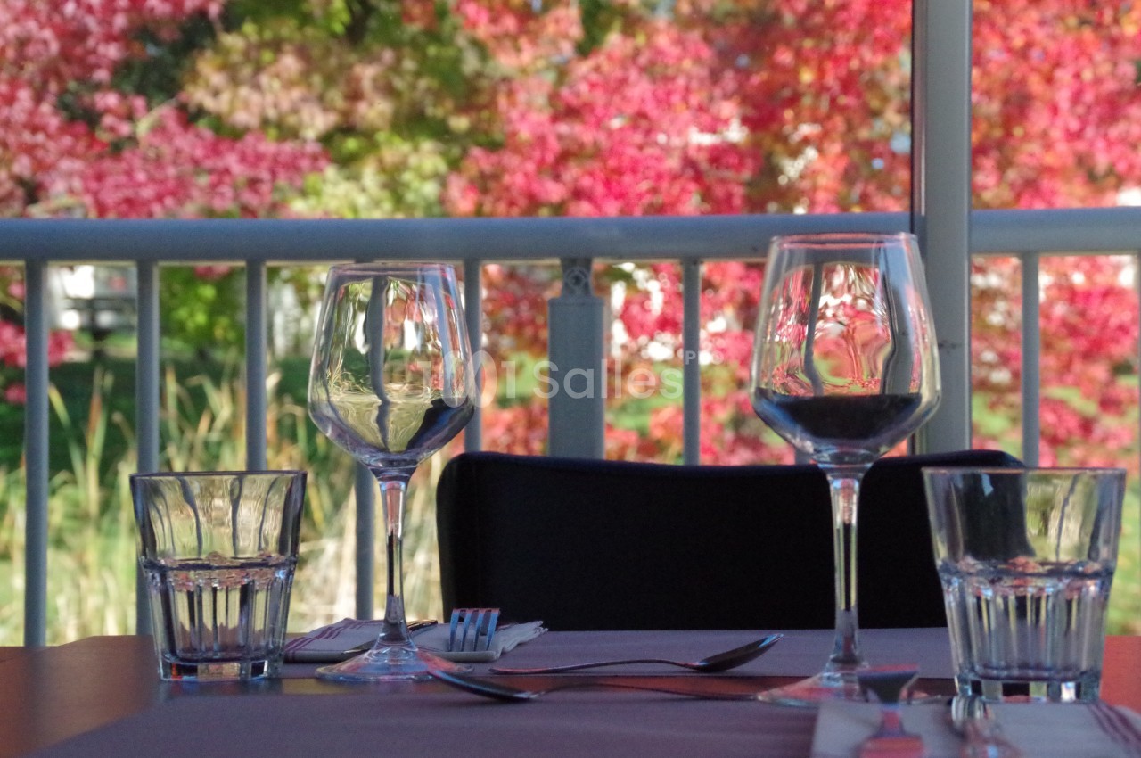 Verres à vin et à eau disposés sur une table, avec des arbres aux feuilles rouges visibles en arrière-plan.