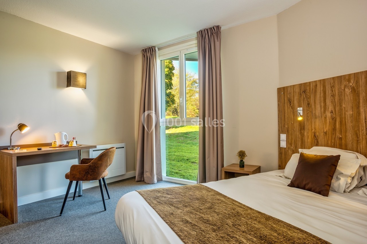 Chambre lumineuse avec lit double, bureau, chaise et fenêtre donnant sur un espace vert.