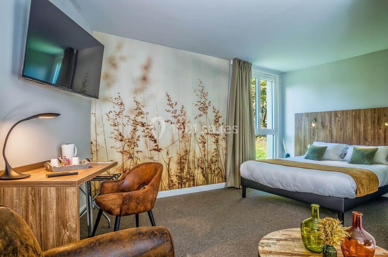 Chambre d'hôtel lumineuse avec lit double, bureau, fauteuils, télévision murale et vue sur un espace vert.
