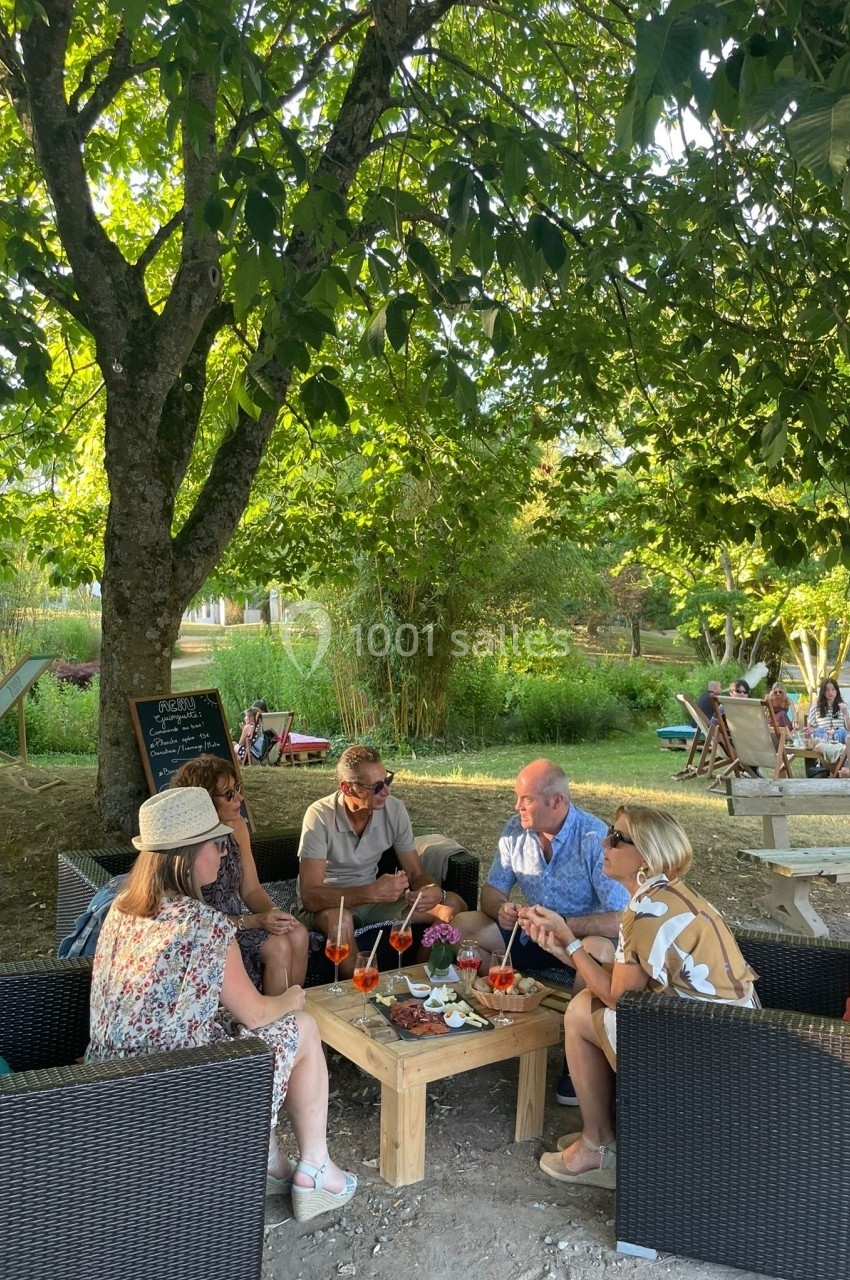 Groupe de personnes assises autour d'une table en plein air, partageant des boissons et des amuse-bouches sous un arbre.