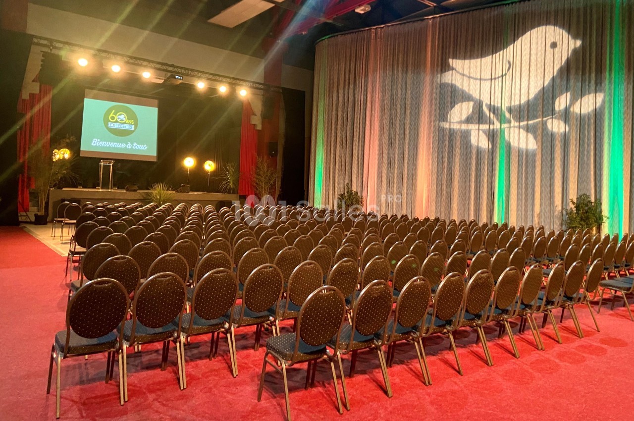 Salle de conférence avec des rangées de chaises vides, une scène éclairée et un écran affichant un logo.