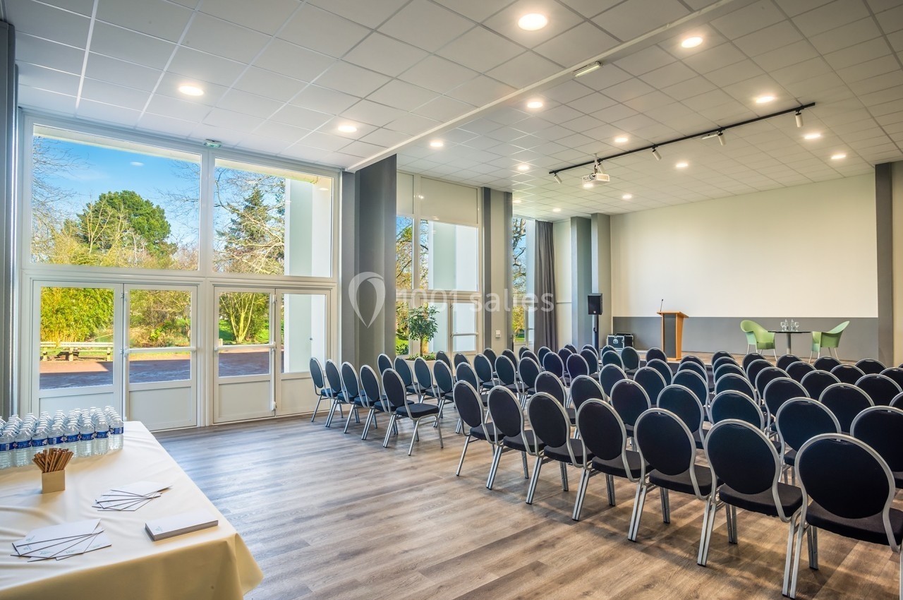 Salle de conférence lumineuse avec rangées de chaises, scène équipée d’un pupitre et grandes baies vitrées donnant sur un…