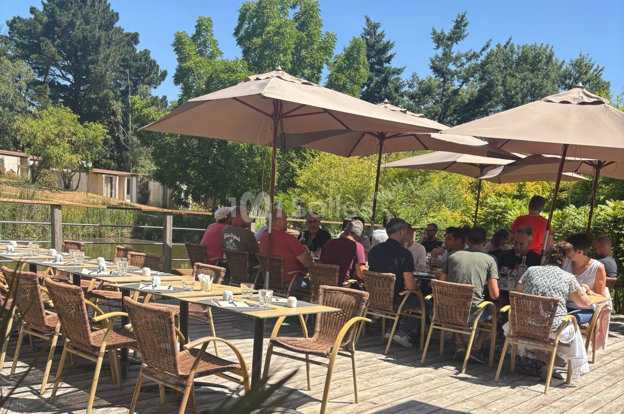 Groupe de personnes assises à des tables en terrasse ombragée, entourée de verdure par une journée ensoleillée.