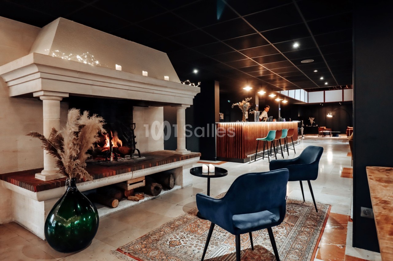 Cheminée allumée dans un espace moderne avec fauteuils bleus, tapis, et un bar éclairé en arrière-plan.