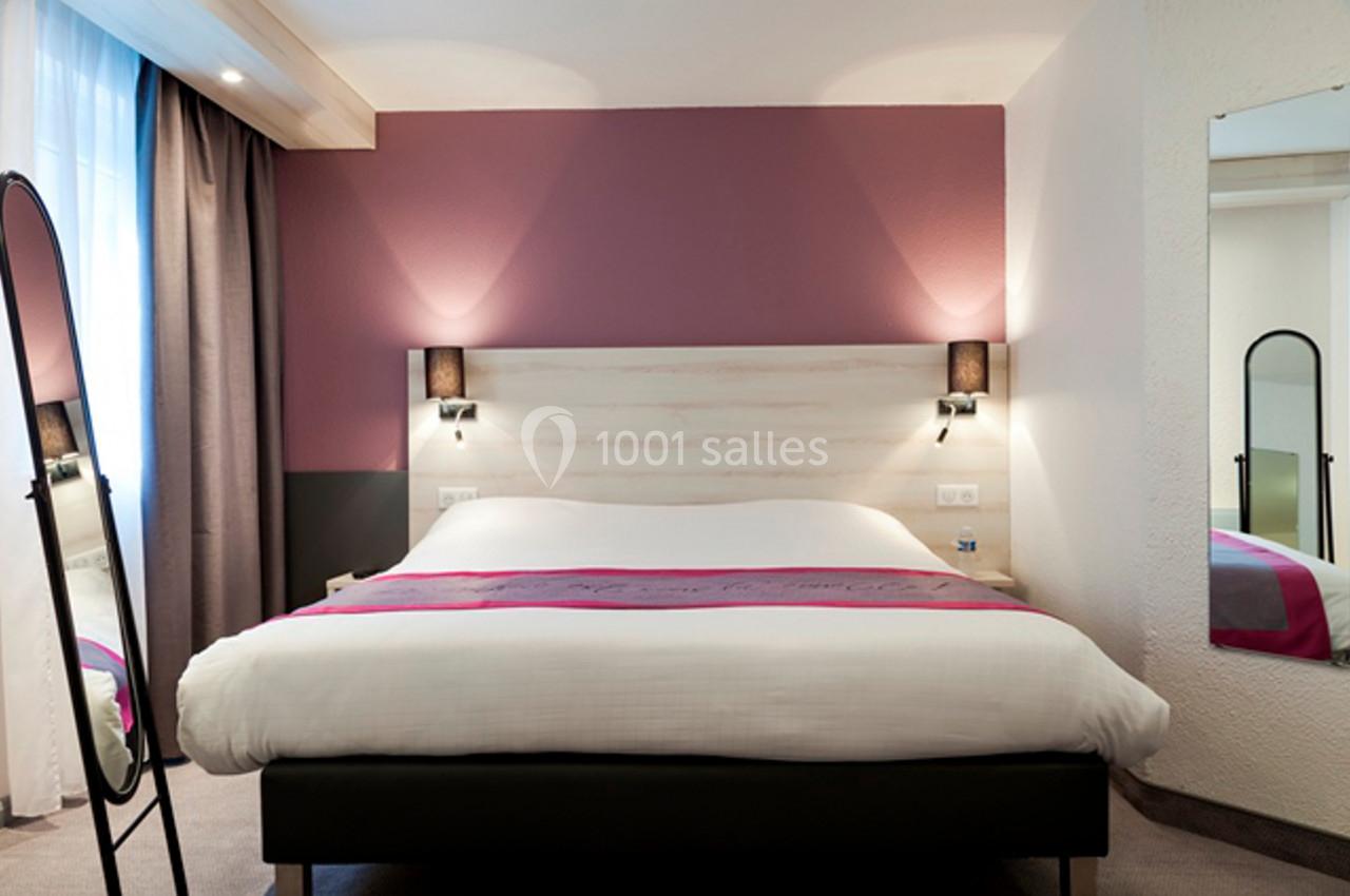 Lit double avec draps blancs dans une chambre moderne aux tons roses et neutres, éclairée par des appliques murales.