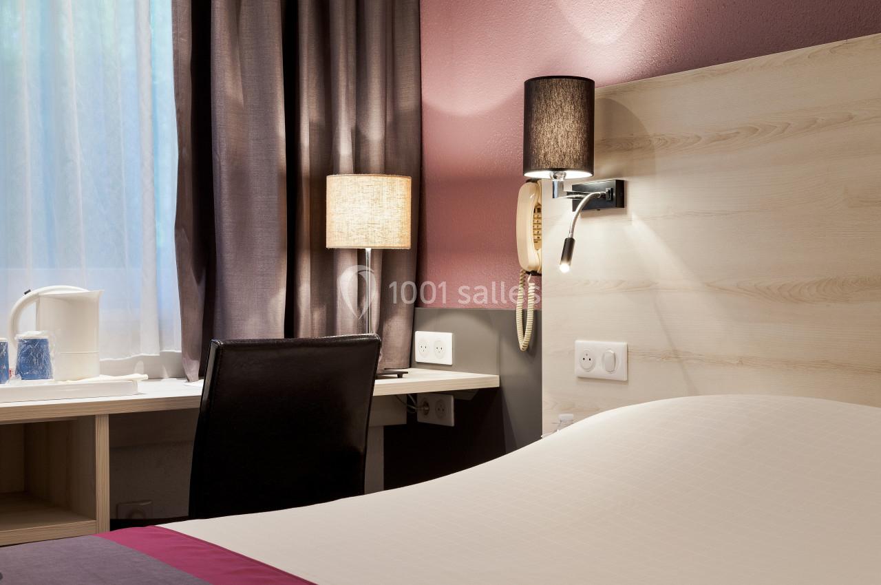 Chambre avec lit, bureau, chaise, lampe de chevet, téléphone mural et lumière naturelle filtrée par un rideau.