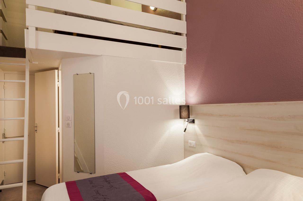 Chambre avec lit double, mur rose, éclairage mural, mezzanine visible en haut et porte ouverte sur un couloir.