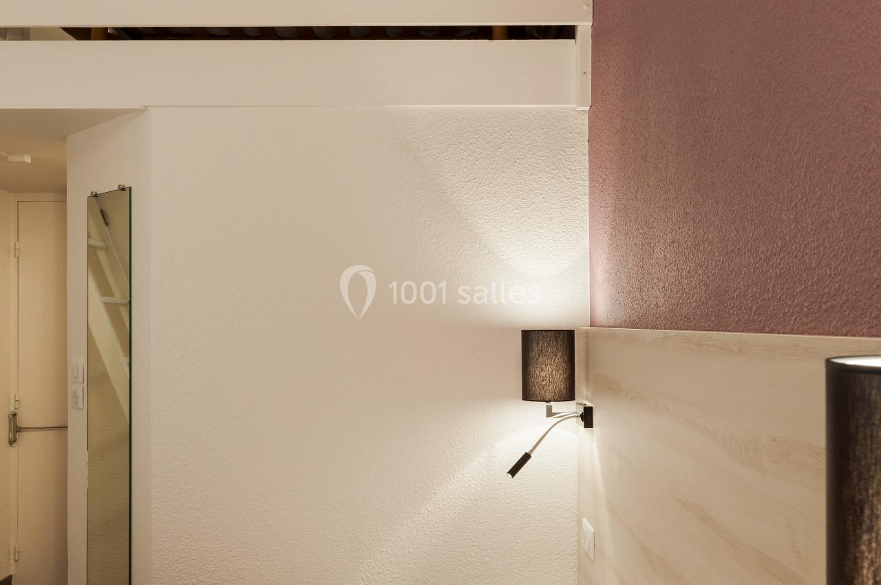 Lit double avec tête de lit en bois clair, éclairage mural et mezzanine visible dans une chambre aux tons neutres.