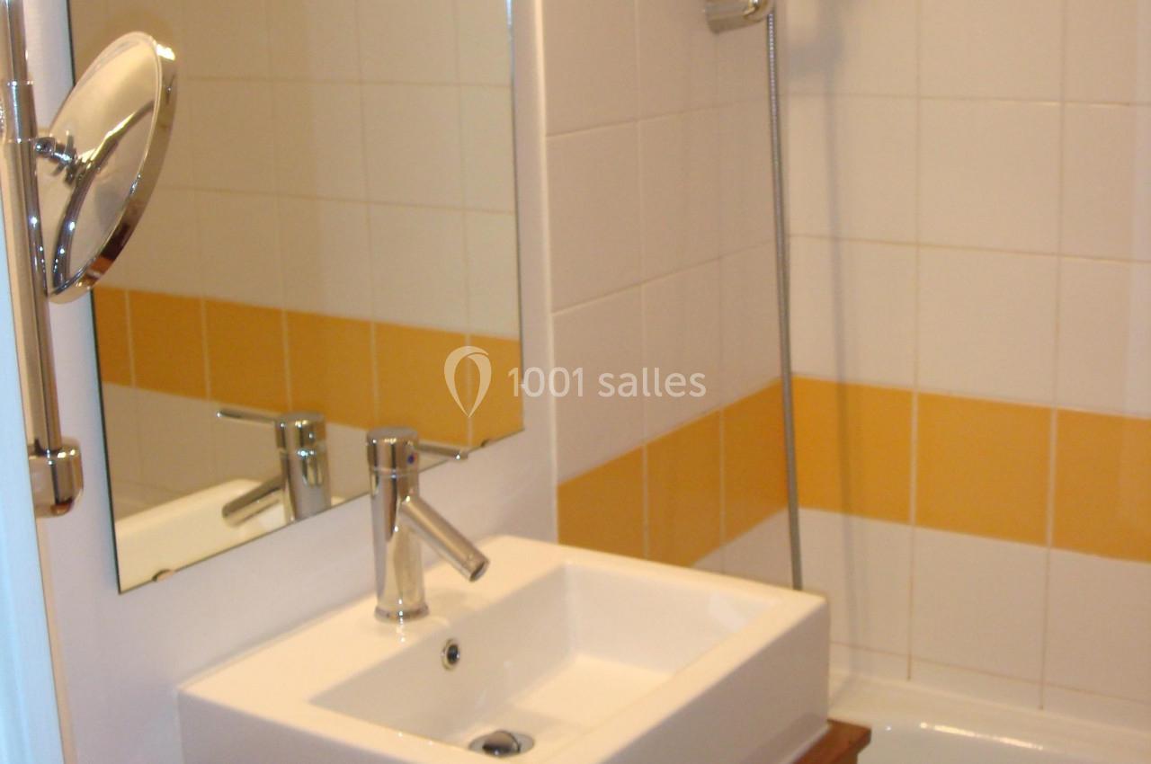 Lavabo blanc sur un plan de travail en bois, avec miroir, douche et carrelage blanc et jaune en arrière-plan.