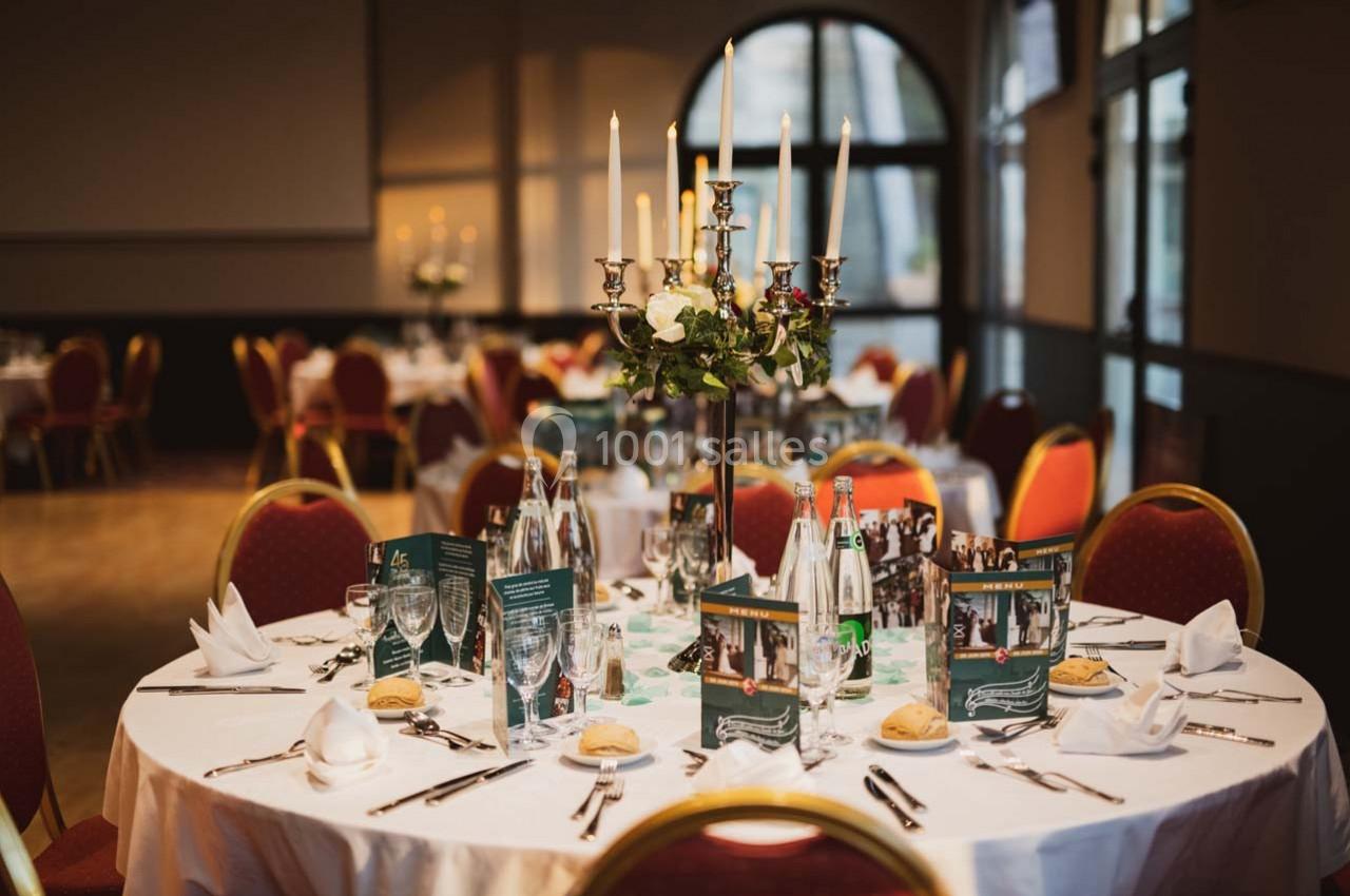 Table dressée avec nappes blanches, chandeliers, menus, verres et couverts dans une salle de réception élégante.