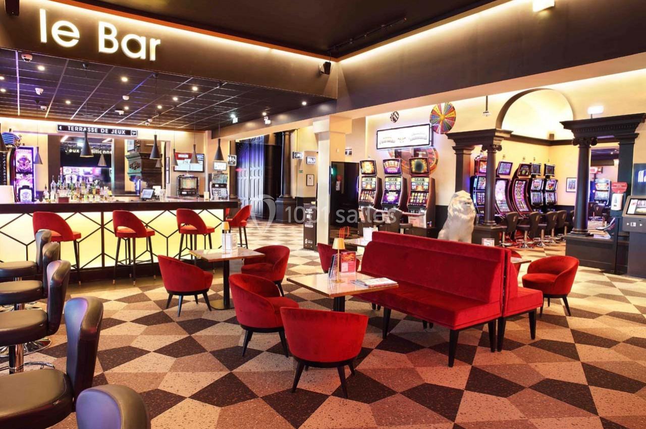 Salle de jeux avec machines à sous, bar éclairé et espace de détente avec fauteuils rouges et tables basses.