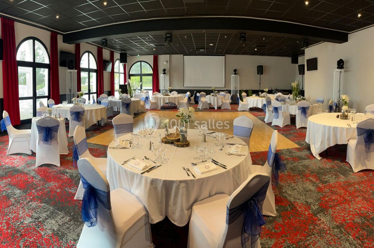 Salle de réception aménagée avec des tables rondes dressées, chaises décorées et écran de projection en fond.